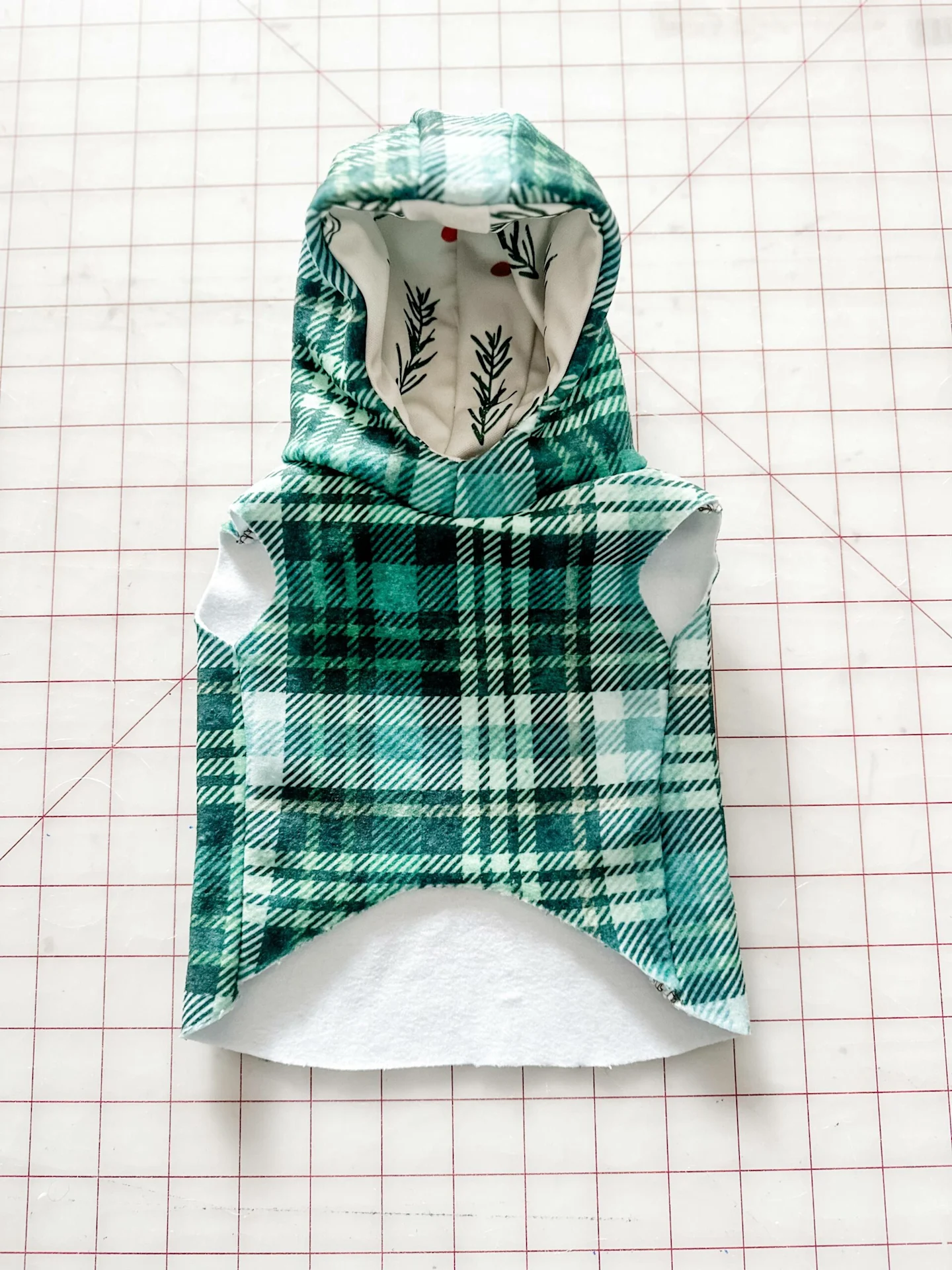 A plaid green-and-white dog jacket is on a white cutting mat with a red grid with the front of the hood facing up. The inside of the hood is white with red dots and evergreen leaves.