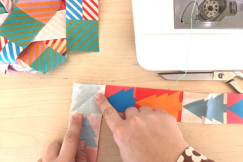A pile of fabric cut in 2” lengthwise strips lay loosely folded on a wooden table. The cotton fabric features a design in jewel-tone colors with small squares of coordinating colors in varying pairings. To the right the bobbin section of a sewing machine 