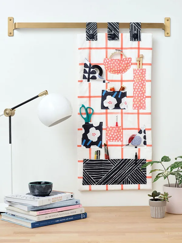 A fabric craft organizer hangs from a wooden dowel attached a wall. It has small pockets, each filled with a different craft tool, including tape, scissors, spools of thread and more. It hangs above a desk with small plants, books and a white lamp.