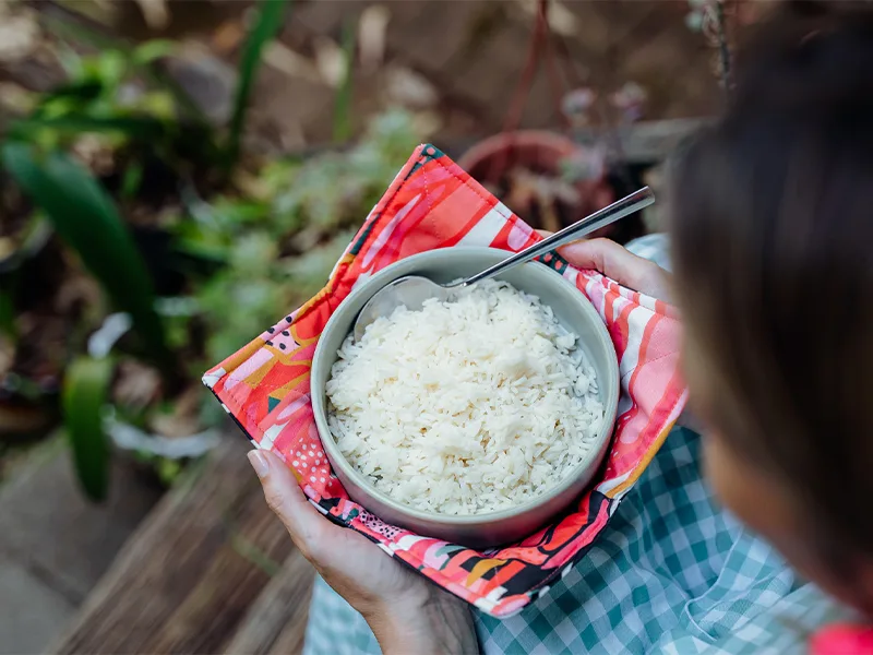 As shown from above and to the left, a person wearing a blue-and-white gingham dress holds a bowl of rice that itself is held in a fabric bowl cozy. The fabric on the top side of the cozy features bright flowers in inks, reds, yellows and white. A spoon i