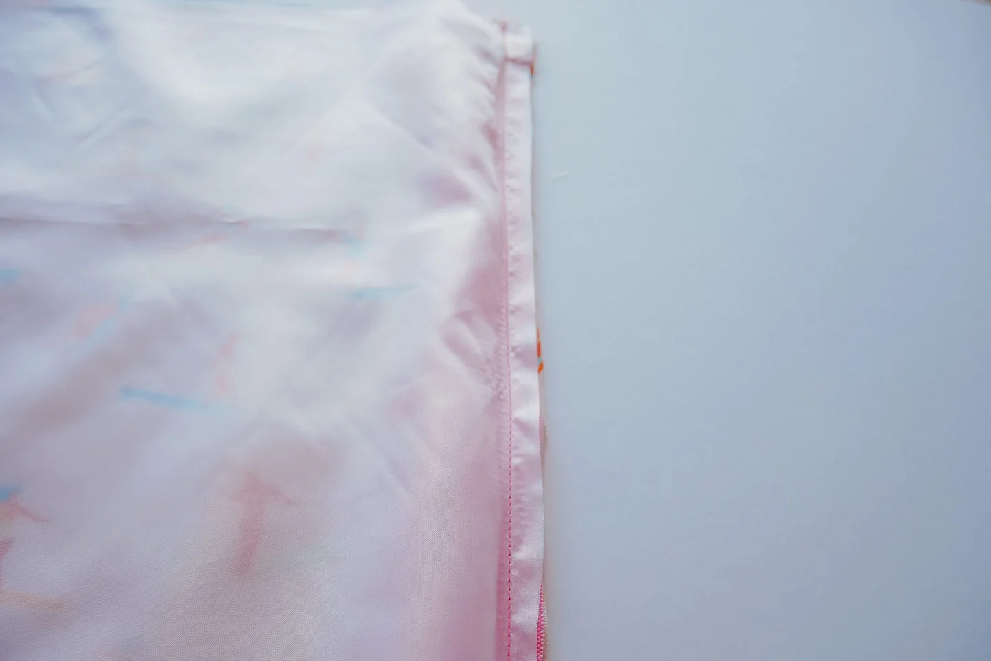 A rectangle piece of Polyurethane Laminate fabric is lying on top of a rectangle of fabric featuring a design with small female surfers surfing through a pink background. A pink zipper lays in between the two. The right edge of those three things has been