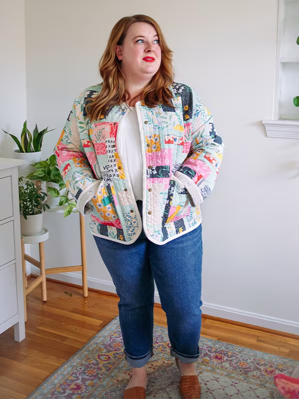 Meg wears her cheater quilt quilted jacket featuring delicate florals, most with a blue or pink theme. She is standing in front of a white wall.