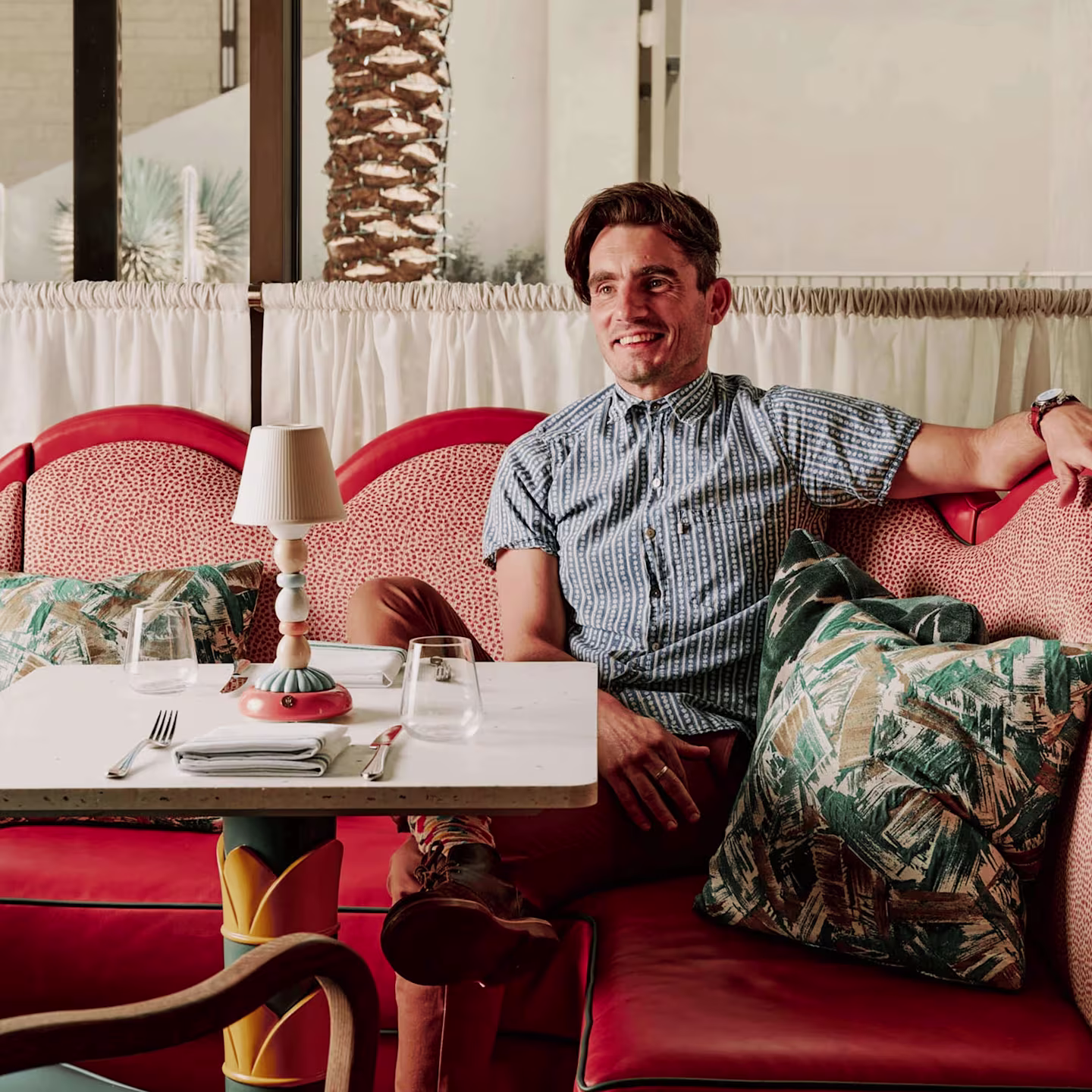 Red upholstered seating with tropical print pillows and layered textiles in a bright restaurant interior with colorful table lamp and natural textures