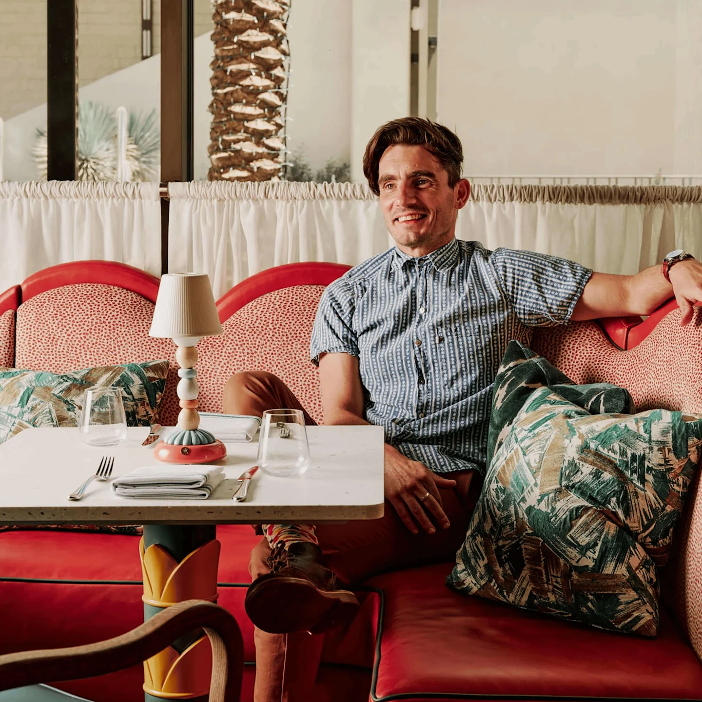 Red upholstered seating with tropical print pillows and layered textiles in a bright restaurant interior with colorful table lamp and natural textures