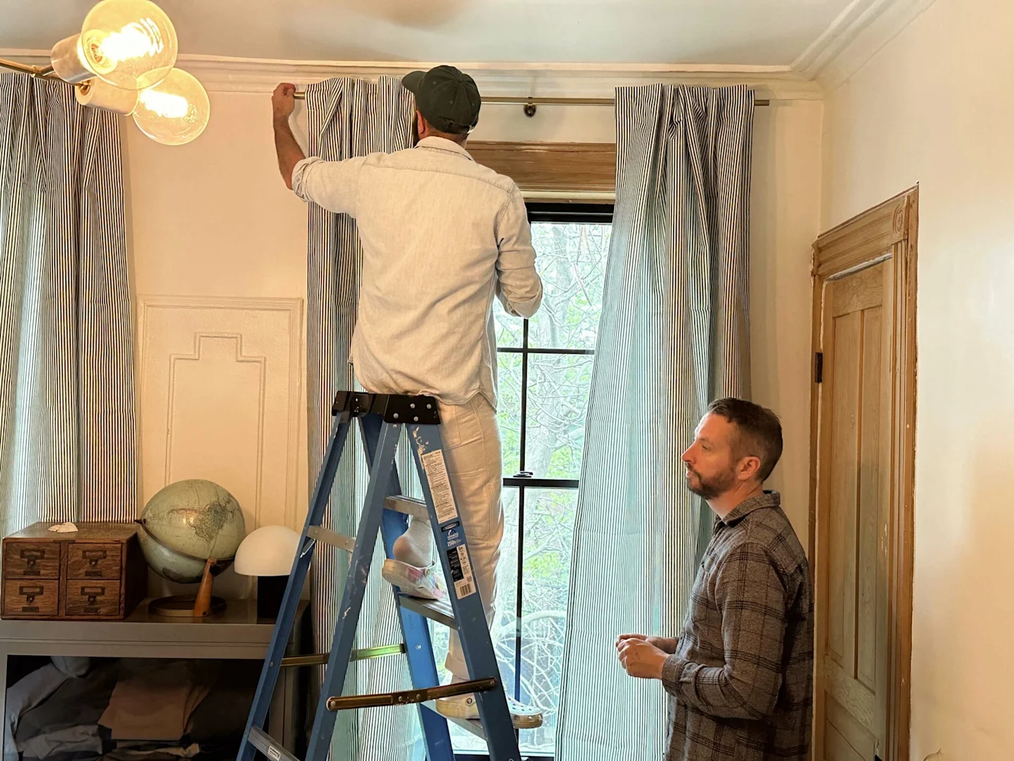 A man stands on a ladder and hangs up the left curtain panel of a pair of top tab dark blue-and-white-vertically striped curtain panels. The right panel has already been hung. Another man stands to the right assisting with the installation. A small bookca