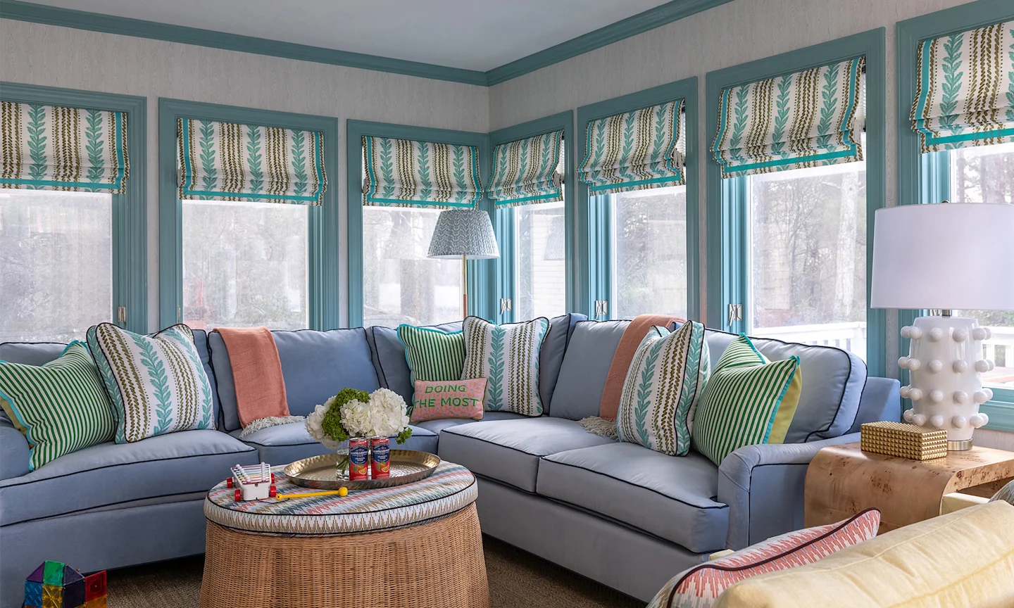 Sunroom with sectional couch thats is covered in throw pillows that match the teal and brown pattern custom roman shades on each window and neutral textured wallpaper.