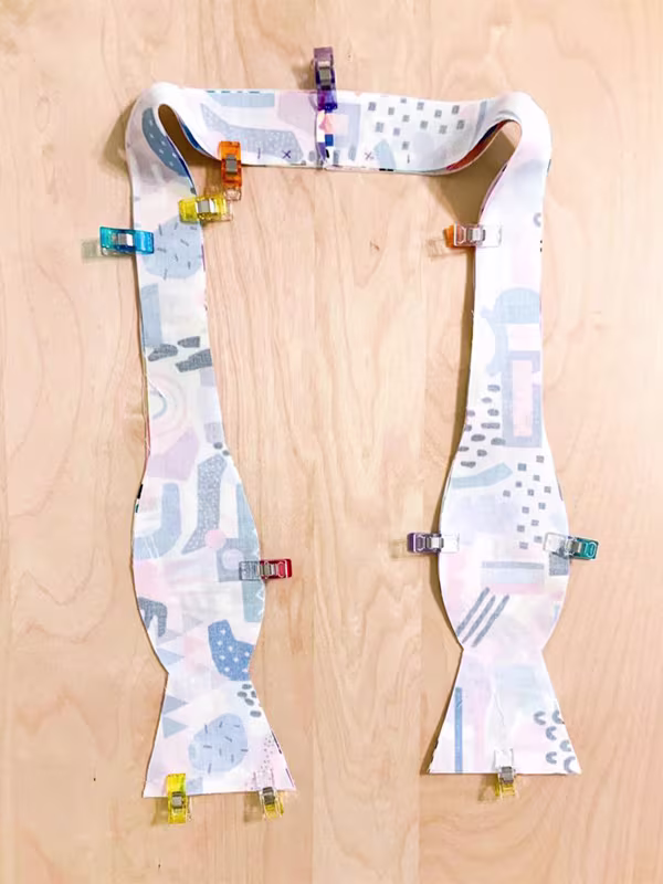 A bowtie laying on a wooden surface design side down and being secured by sewing clips.