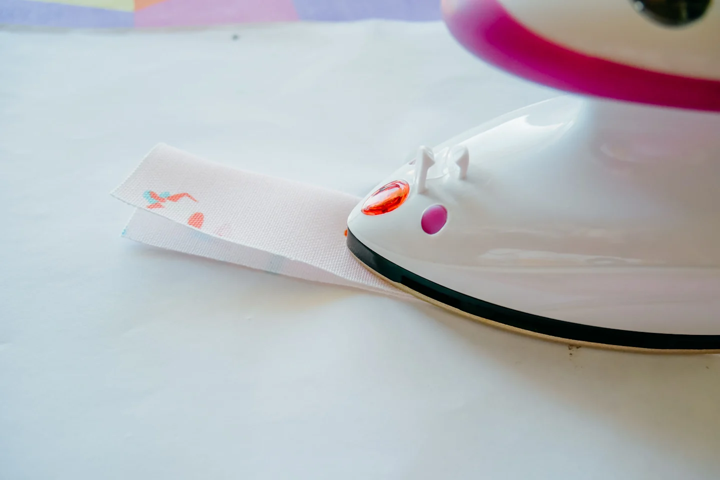 A small rectangle of fabric has been folded in half lengthwise with the wrong sides of the fabric together and is being pressed together with a hot iron. The fabric features a design with small female surfers surfing through a pink background.