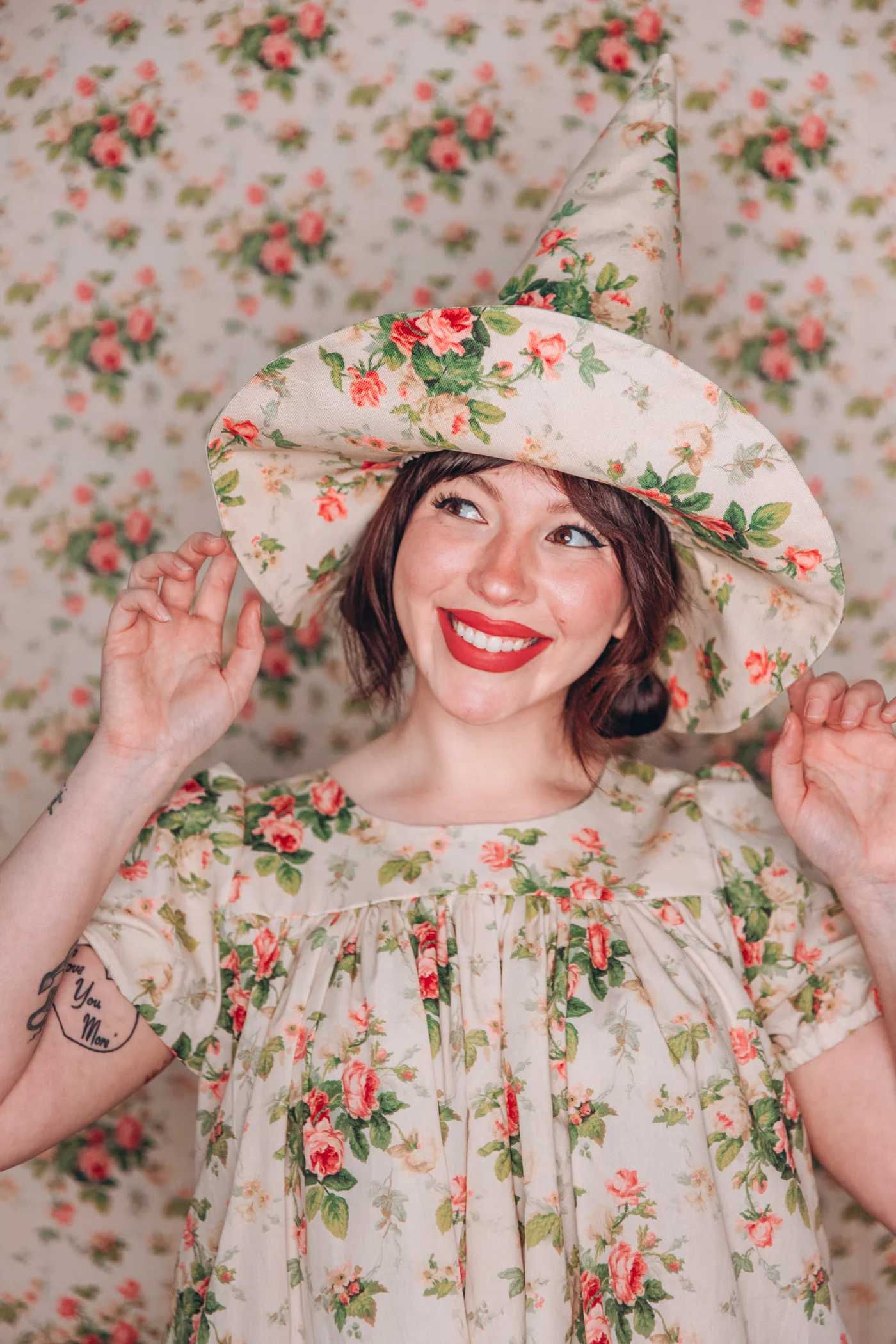 Keiko wearing a floral witch hat in a matching dress in front of a backdrop of the same design. She's smiling with the tips of her fingers touching the brim of her hat.