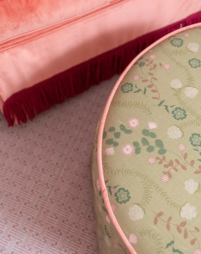 Round floral upholstered ottoman beside a coral armchair with fringe trim on a pale pink patterned rug.