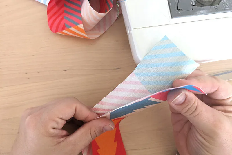 A 2”-wide curled up strip of fabric with a design featuring small squares of coordinating colors in varying pairings is to the top left. Two hands are lining up two 2”-wide strips of fabric at the tops, wrong sides together, in the middle of the image. Th