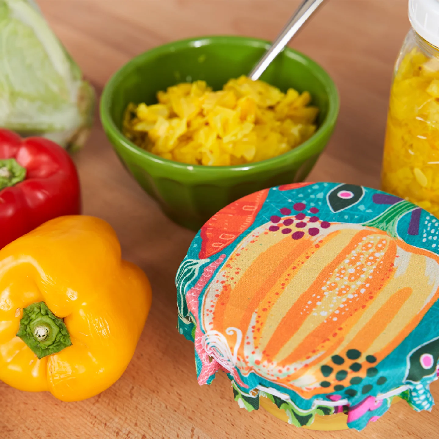 A completed no-sew bowl fabric cover sits on top of a small bowl to the photo’s bottom right. To the top right of the photo is a bowl of homemade chow chow in a green bowl. A head of cabbage, a red pepper, a yellow pepper and a yellow squash are to the le