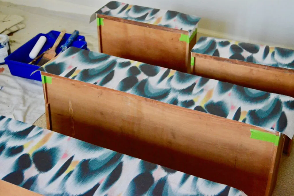 Four dresser drawers lay facing upward with measured pieces of wallpaper laying on top of them. Other materials sit in the distance.