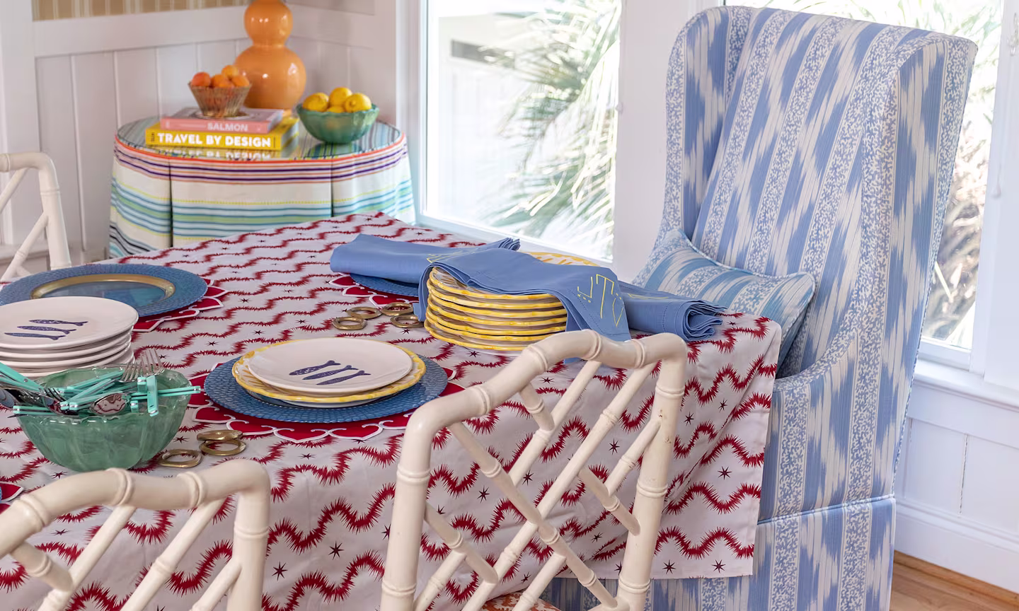 A vibrant, coastal-inspired dining room featuring a bold patterned tablecloth, blue striped seating, colorful tableware, and fresh citrus accents bathed in natural light.