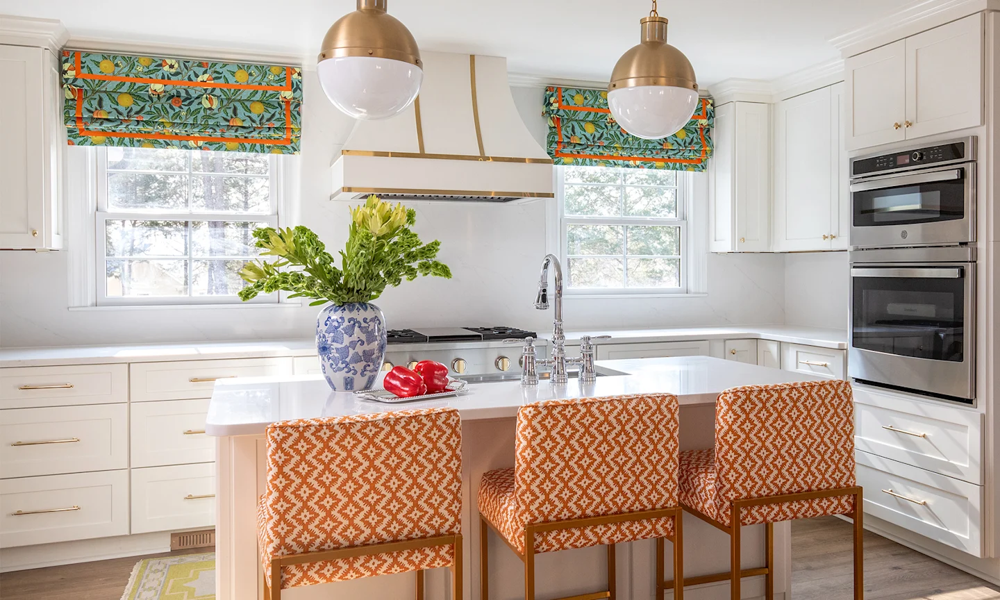 Kitchen area with island and chairs upholstered in a geometric orange fabric and roman shade in a william morris inspired fruit print.