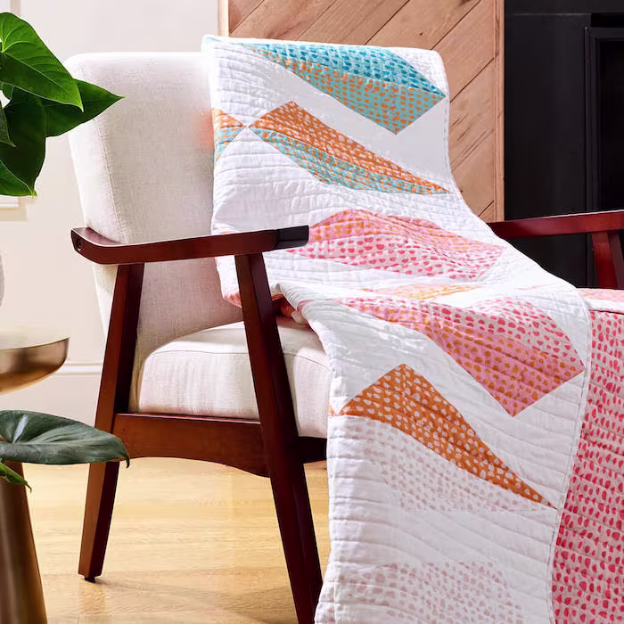 A modern armchair with a white cushion has a quilt draped over it. The quilt features geometric shapes in bright colors, including orange, blue, and pink, arranged in angled, overlapping sections.