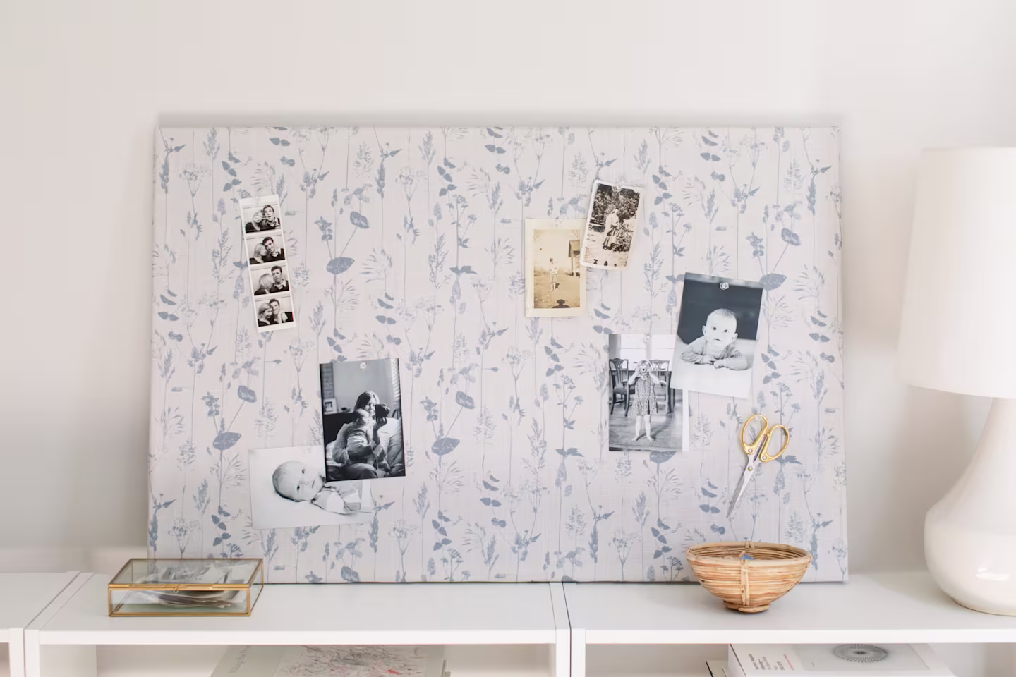 A close up of the bulletin board’s fabric cover. The fabric design has a white background and light blue flowers and the weave of the fabric is highlighted at this distance. Two black-and-white vintage photos, one of a woman holding a baby in front of a h