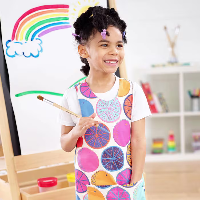A child holding a paintbrush stands in front of an easel with a colorful rainbow painting. The child is wearing a paint smock decorated with large, circular, multicolored fruit-inspired patterns.