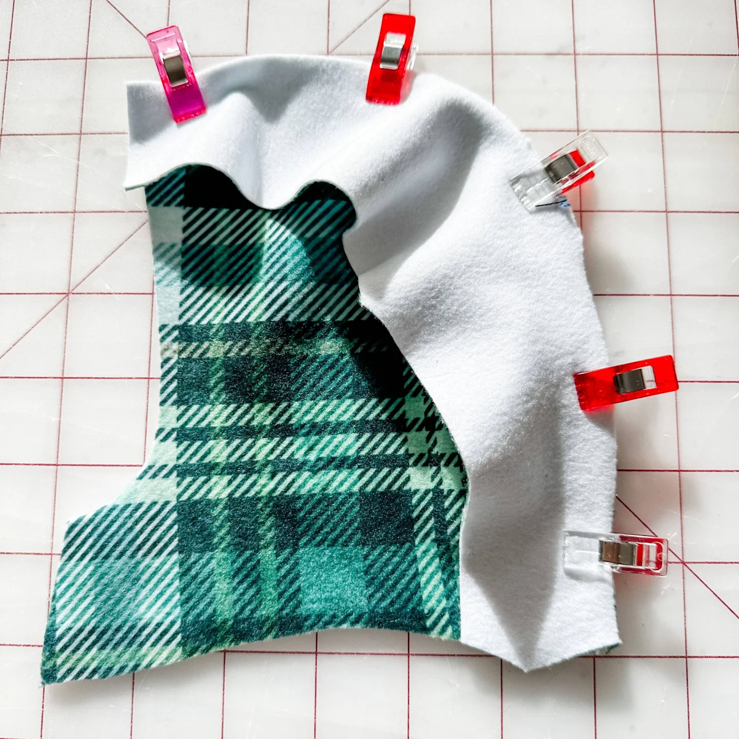A side piece of the hood is design side up and is clipped to the center piece using sewing clips. The hood’s center piece is design side down. The fabric lays on a white cutting mat with a red grid. The design on the fabric is a green-and-white plaid.