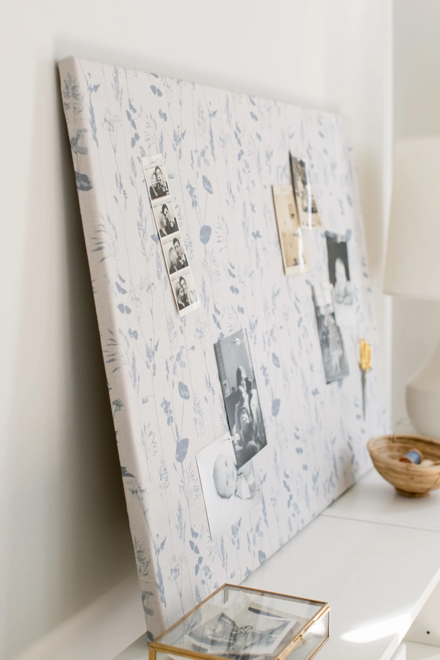 A close up of a fabric-covered bulletin board resting on the top of a low white bookshelf. The fabric design has a white background and light blue flowers. To the board’s bottom right are two black-and-white photos of small children and a pair of scissors