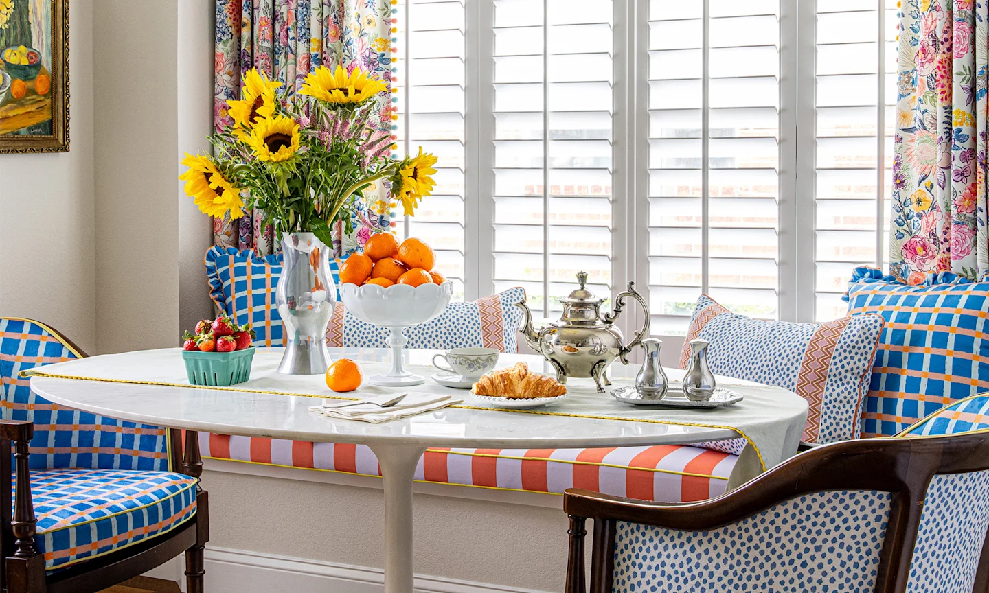 Dining area with blue checkered chairs, pillows an orange and pink striped window seat and floral curtains.