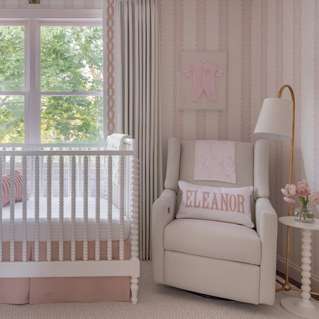 Soft pink nursery with striped wallpaper, white crib, upholstered glider, personalized pillow, and floral accents.