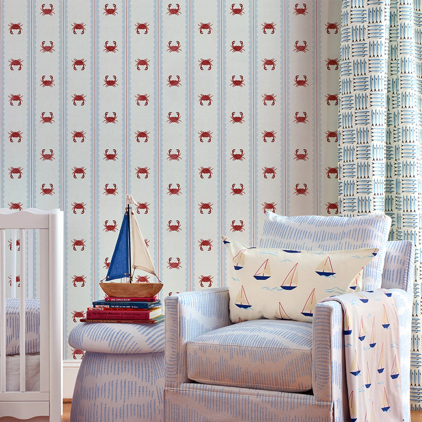 Nursery with striped crab wallpaper, blue upholstered chair and ottoman, sailboat pillow and blanket and sardine curtains.