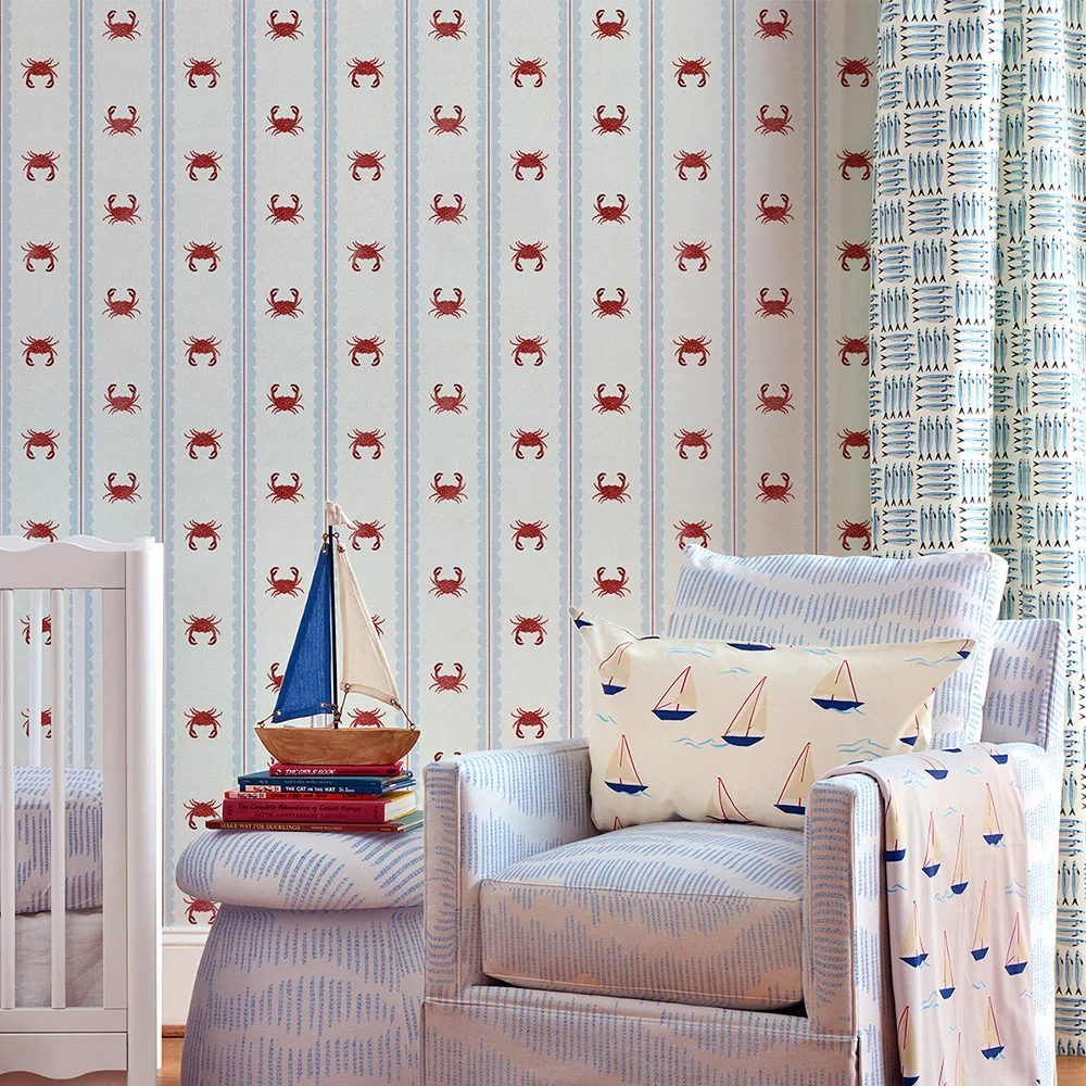 Nursery with striped crab wallpaper, blue upholstered chair and ottoman, sailboat pillow and blanket and sardine curtains.