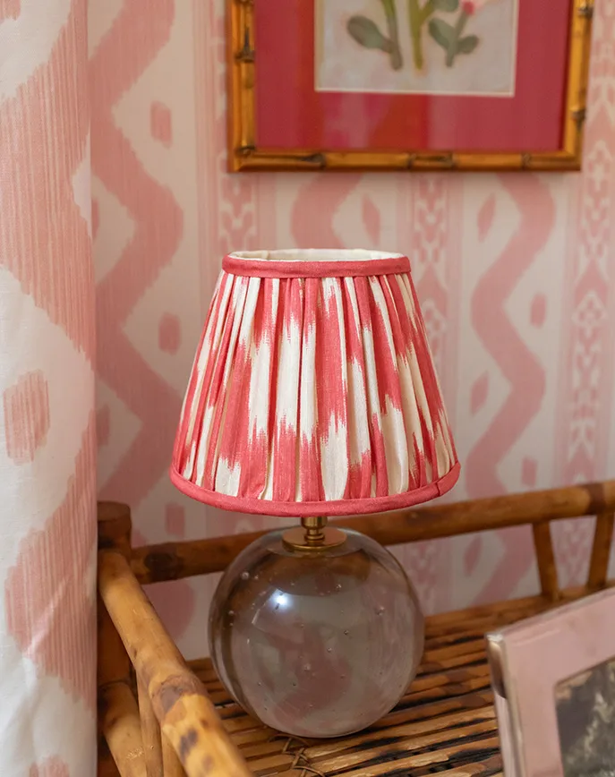 Pink and white patterned wallpaper displayed behind a wooden bookshelf with a pleated table lamp and framed artwork.