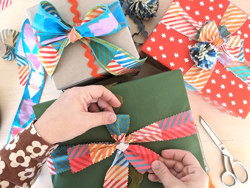 Three presents are on a table, all wrapped with fabric ribbons tied in bows. Two hands are tying the bow on top the present toward the bottom middle of the image. The bottom middle and right present are tied with fabric ribbon with a design featuring smal
