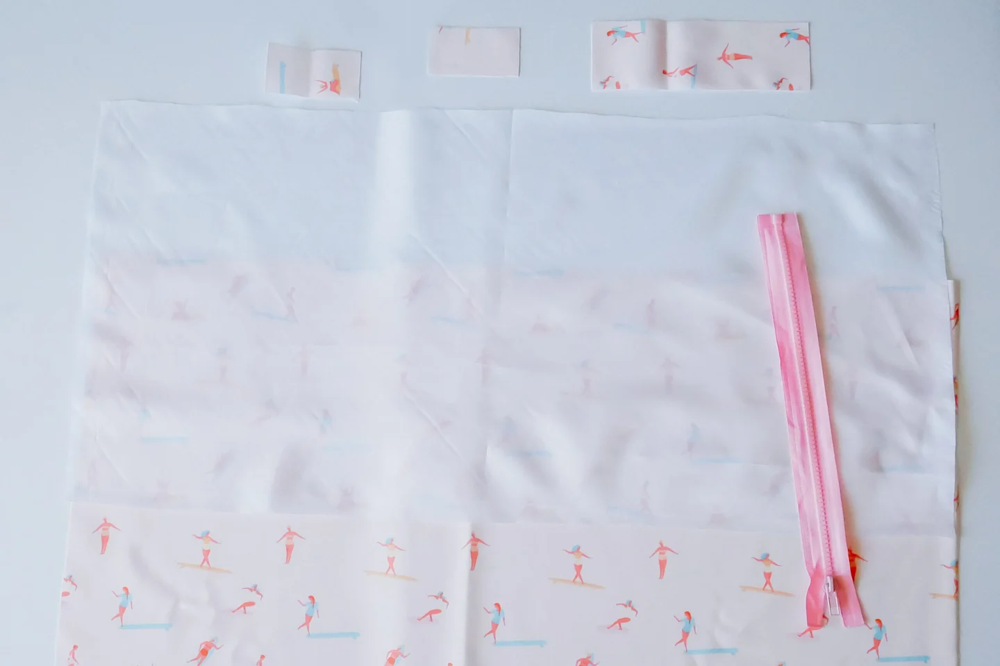 Two rectangle pieces of fabric for the wet bag are lying on a white surface. The piece on top has been cut out of Polyurethane Laminate. The piece on the bottom has a design featuring small female surfers surfing through a pink background. Three smaller r