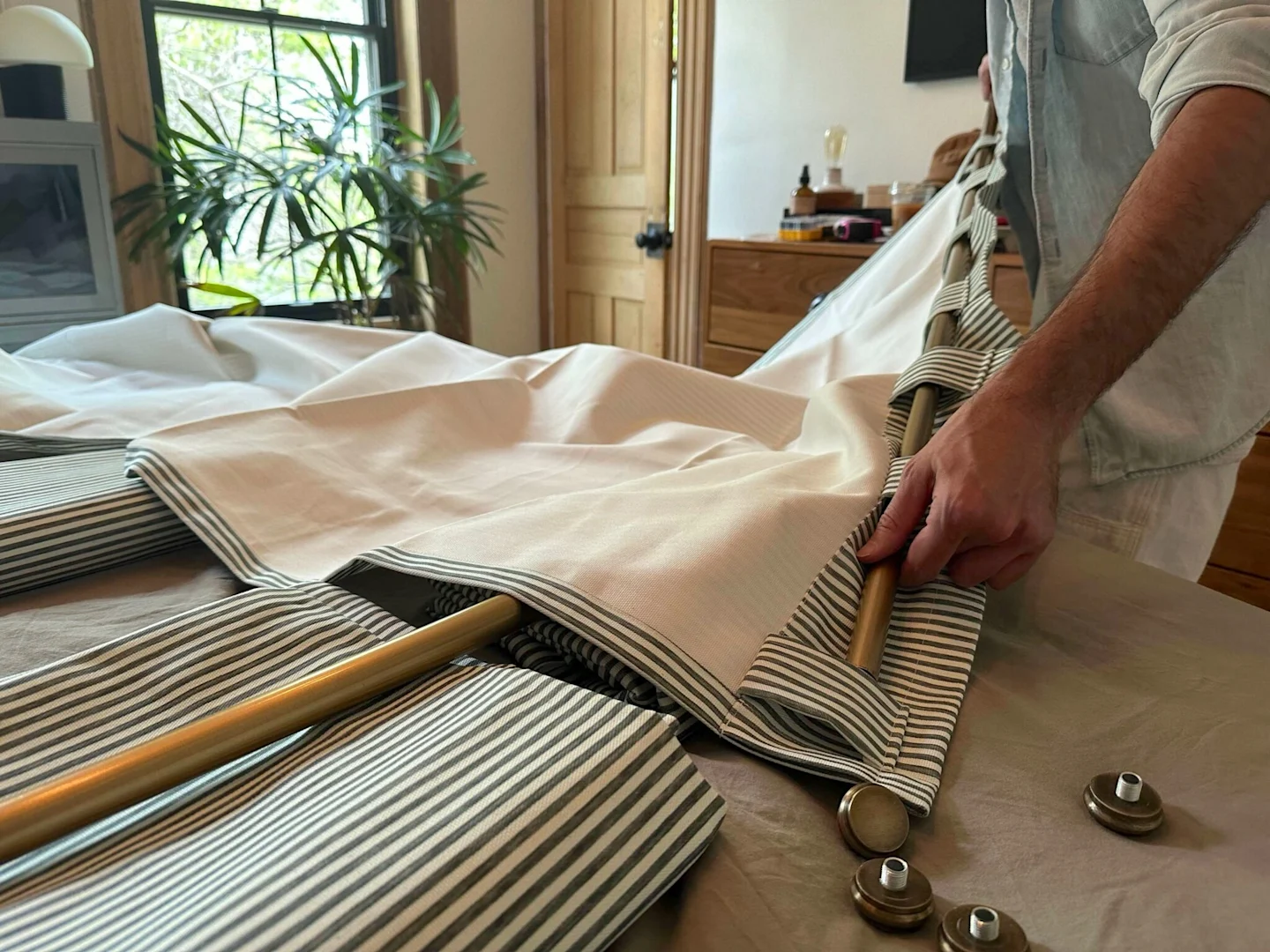 A man is sliding a metal curtain rod through the top tabs of a dark blue-and-white-vertically striped curtain panel. Only his hands are shown as he guides the curtain rod through the fabric. A wooden door is at the top of the image and a large window and 