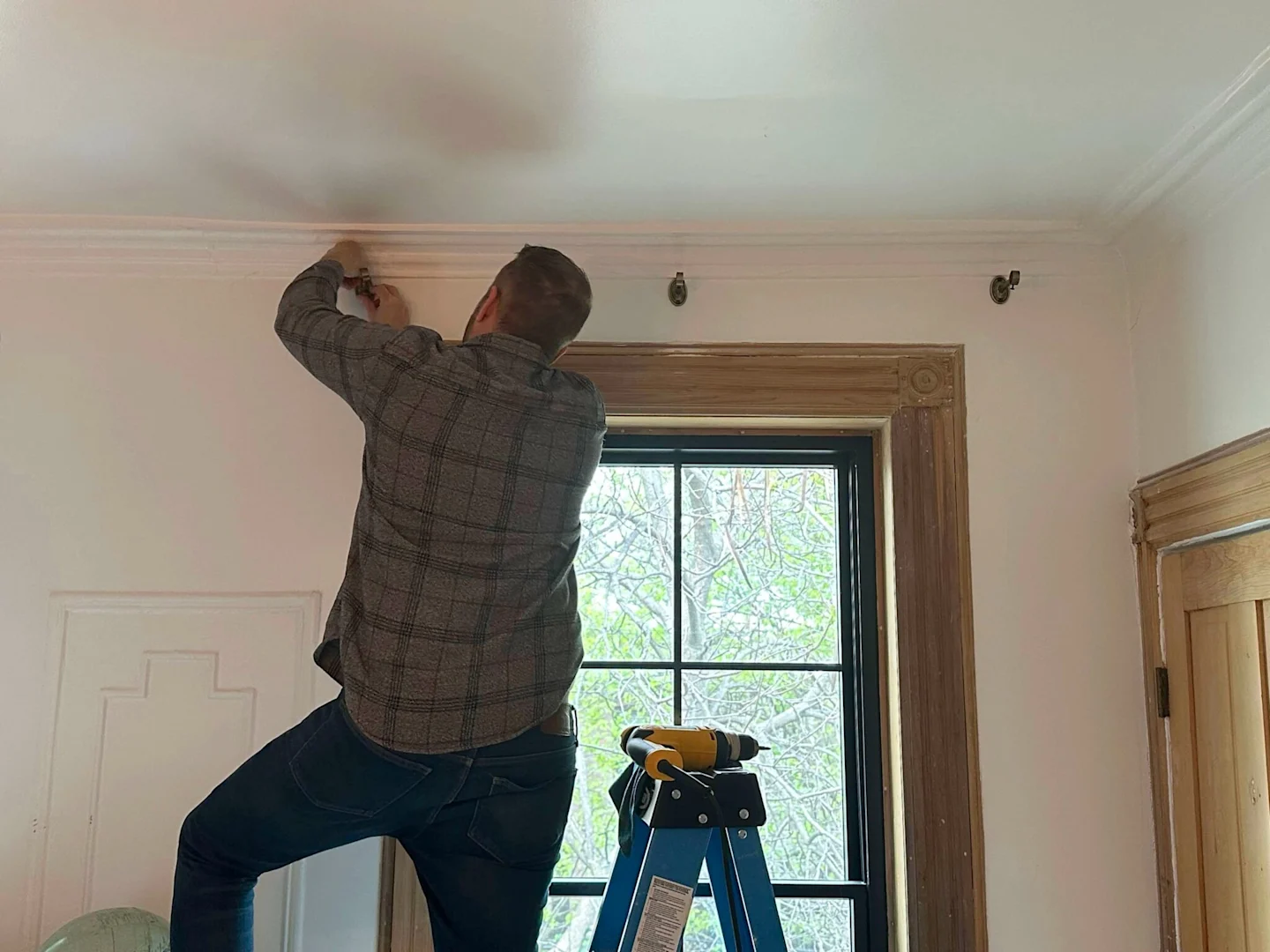 A man installs metal curtain brackets above a window with brown wooden molding. He is standing on a ladder.