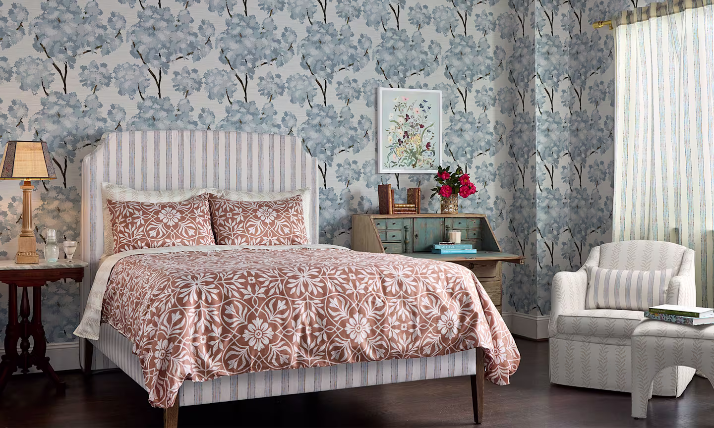 Bedroom with blue tree wallpaper, terracotta medallion bedding, striped curtains and upholstered headboard and chair.