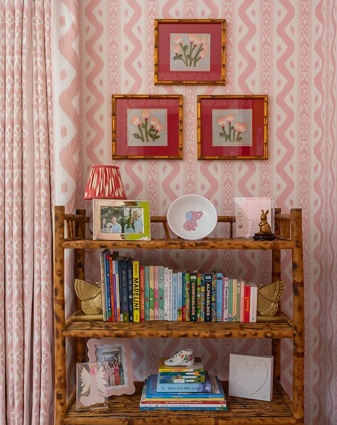 Pink patterned wallpaper behind a wooden bookshelf styled with children's books, framed artwork, and decorative accents.