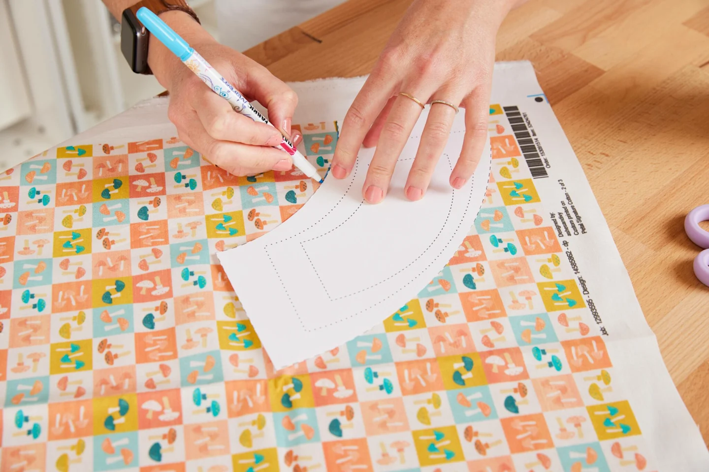 Alexa traces the cut-out pattern piece for a 4” (10.2 cm) flower pot from our tutorial template onto fabric that features small squares each featuring small mushrooms in blue, white, green and peach is laying on the table.