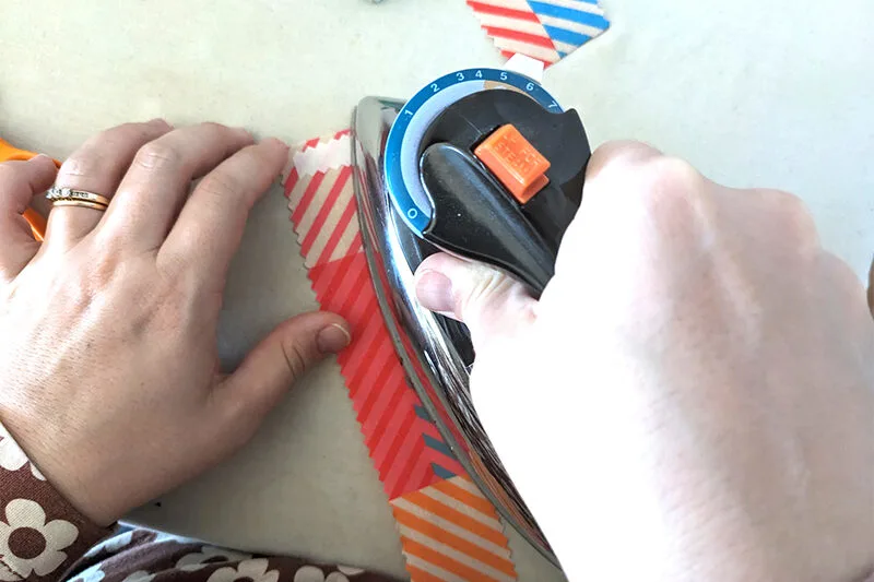 An iron is adhering iron-on adhesive to a long strip of 2”-wide cotton fabric featuring a design with small squares of coordinating colors in varying jewel tone pairings has been cut out with pinking shears. The fabric is laying on a white surface.