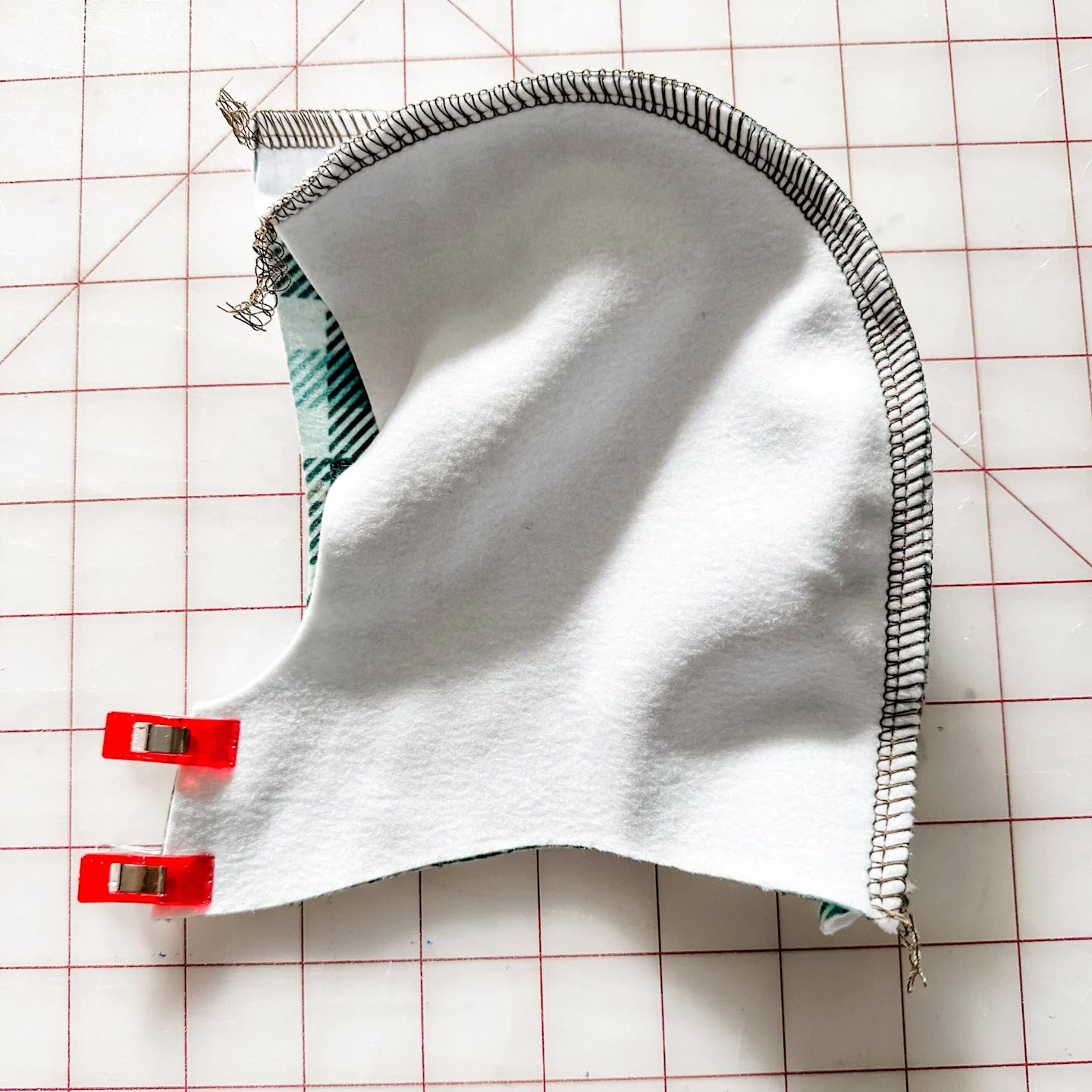 One of the hoodie’s side pieces is shown design side down and sewn to the curve of the center piece. Two sewing clips are at the hoodie’s small front center piece holding the two side pieces together. The fabric lays on a white cutting mat with a red grid