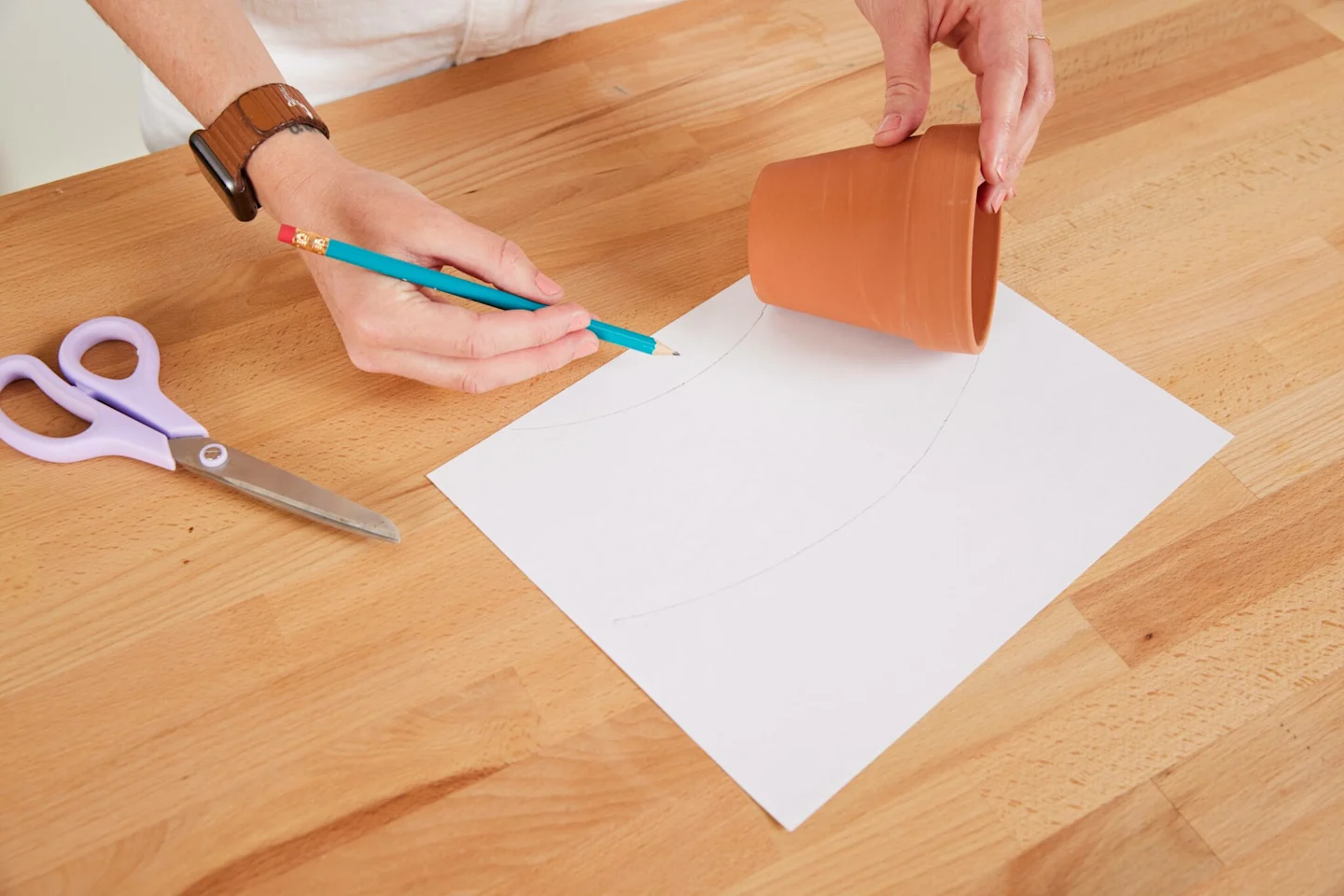 Alexa uses a pencil and a piece of paper to create a paper template for her fabric-covered flower pot. She has rolled the surface of the flower pot lengthwise across the paper from left to right, tracing the edge of the flower pot as she goes.