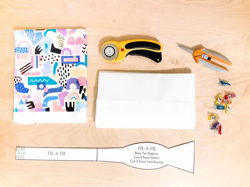Materials to make a bow tie including fabric, a rotary blade, clips, scissors, interfacing and template.