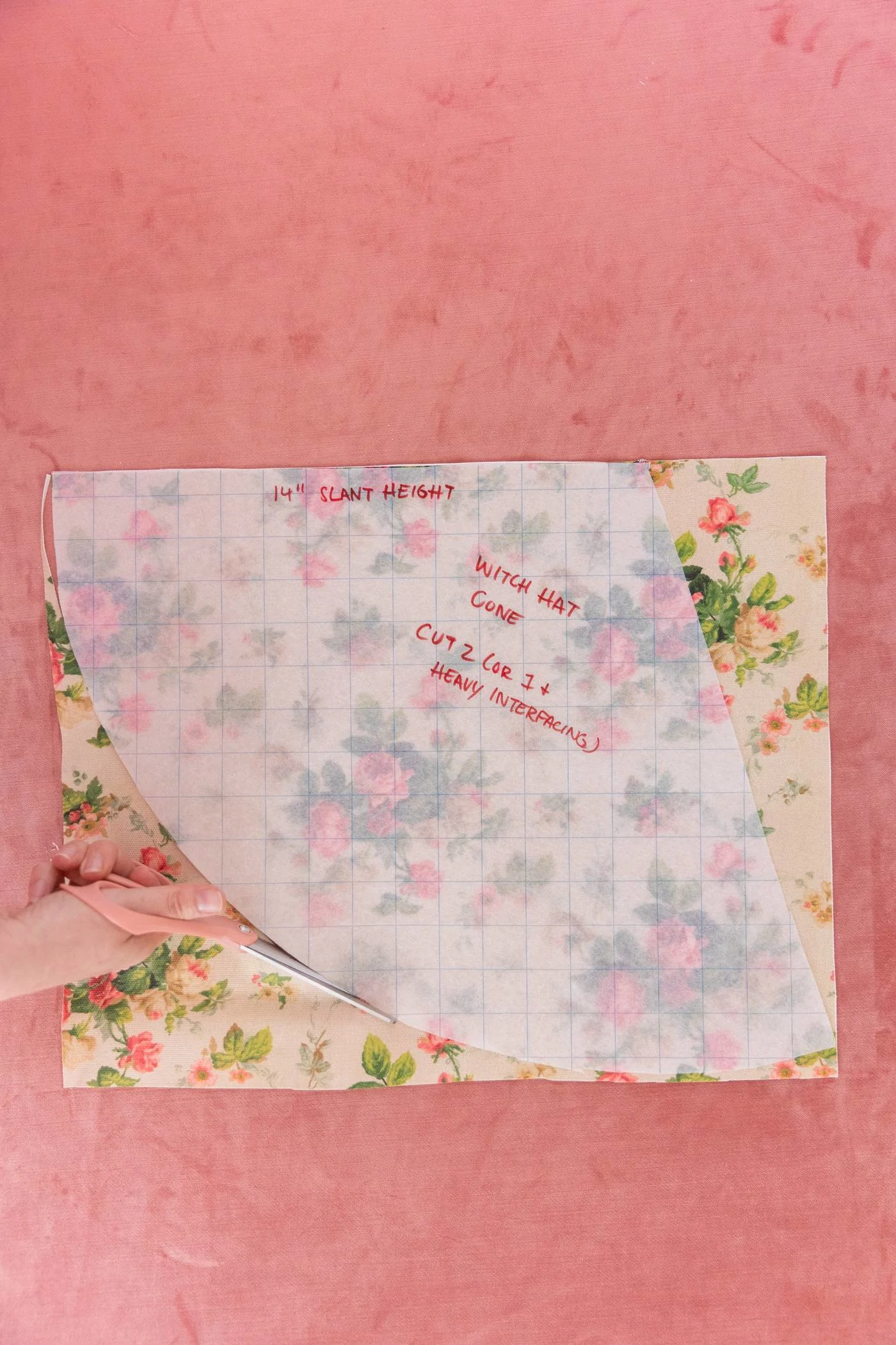 Keiko cuts a cream fabric with pink roses and green leaves in the cone shape of the translucent grid paper resting on top of the fabric, with a peach-colored pair of scissors. A pink carpet is in the background.