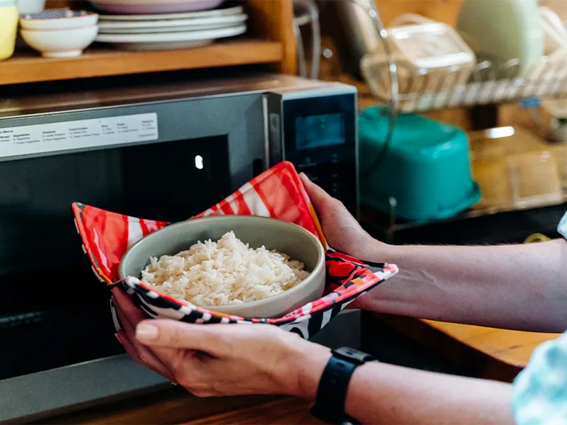 A person wearing a blue-and-white gingham dress puts a bowl of rice that itself is held in a fabric bowl cozy in the microwave. The fabric on the top side of the cozy features bright flowers in inks, reds, yellows and white. The fabric on the bottom side 