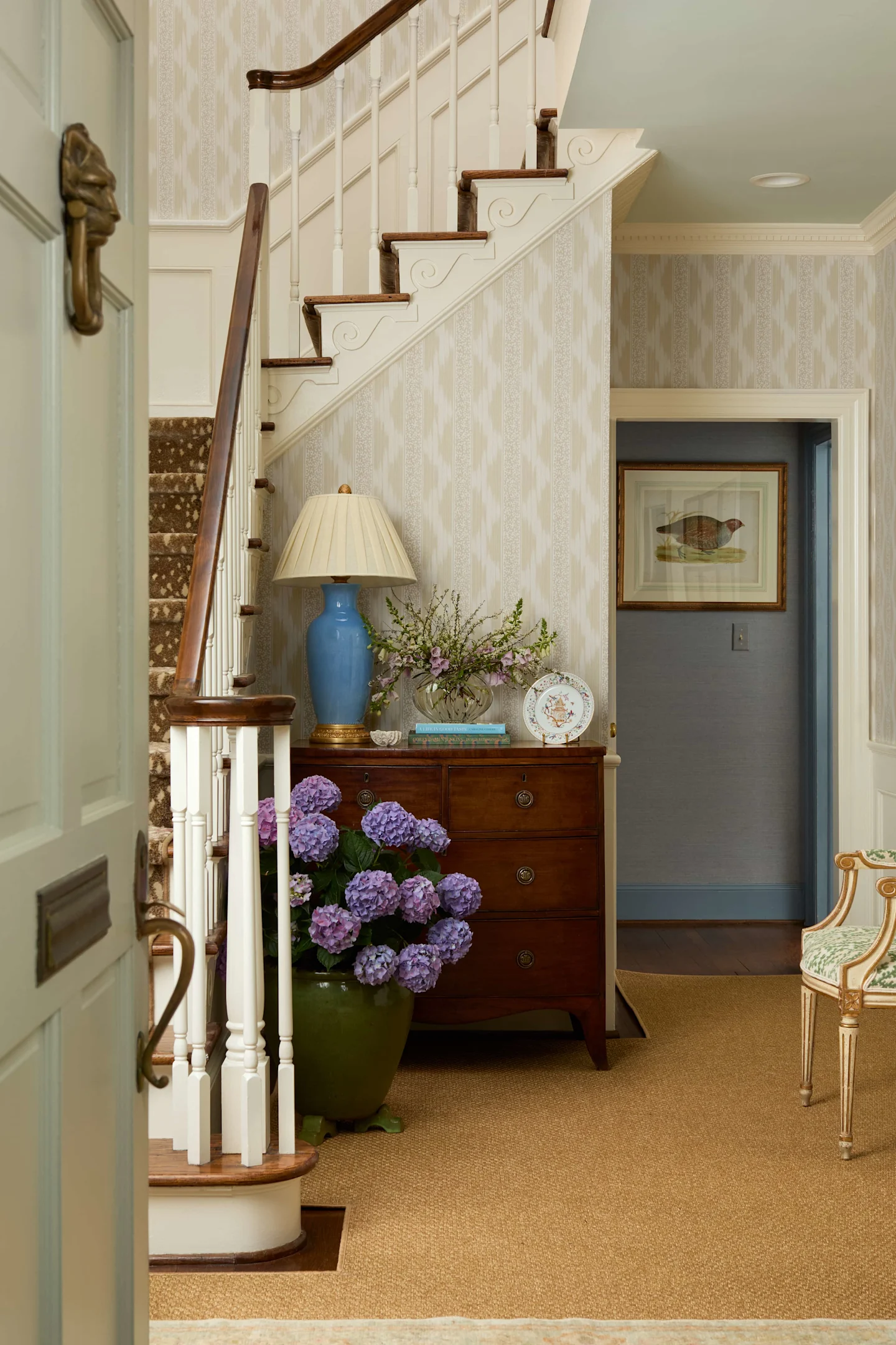 Neutral patterned wallpapered entryway with white staircase and wood railing, wooden chest with lamp and floral arrangement, and hydrangea plant in ceramic pot.