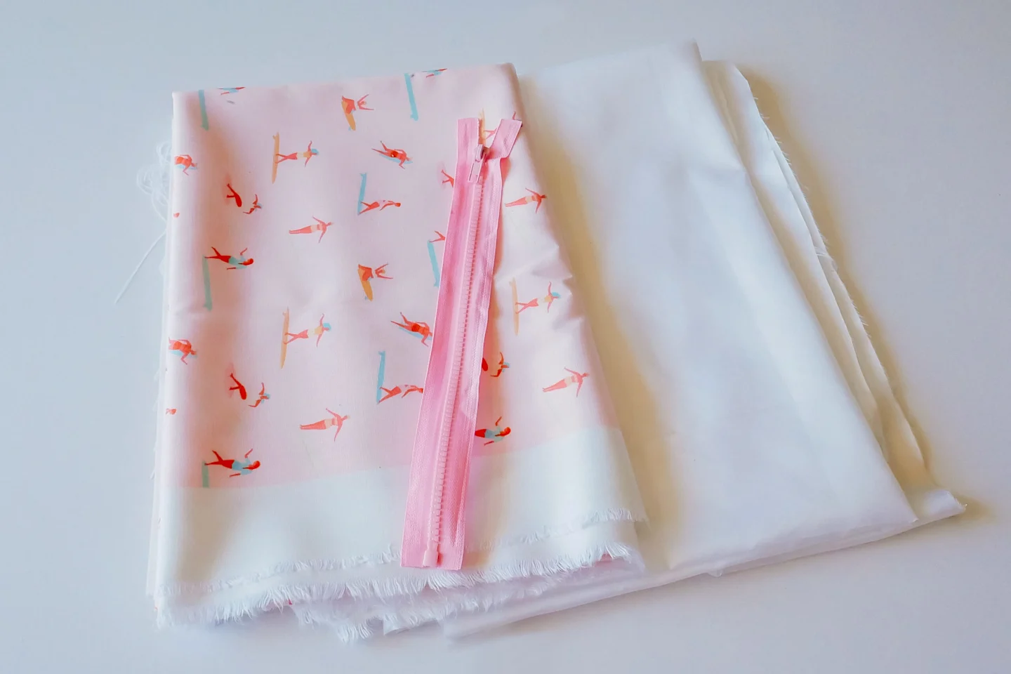 A yard of recycled canvas fabric in a design with small female surfers surfing through a pink background is folded and lying on top of a yard of folded Polyurethane Laminate for the outside and inside of the wet bag, respectively. A 10” (25 cm) pink zippe