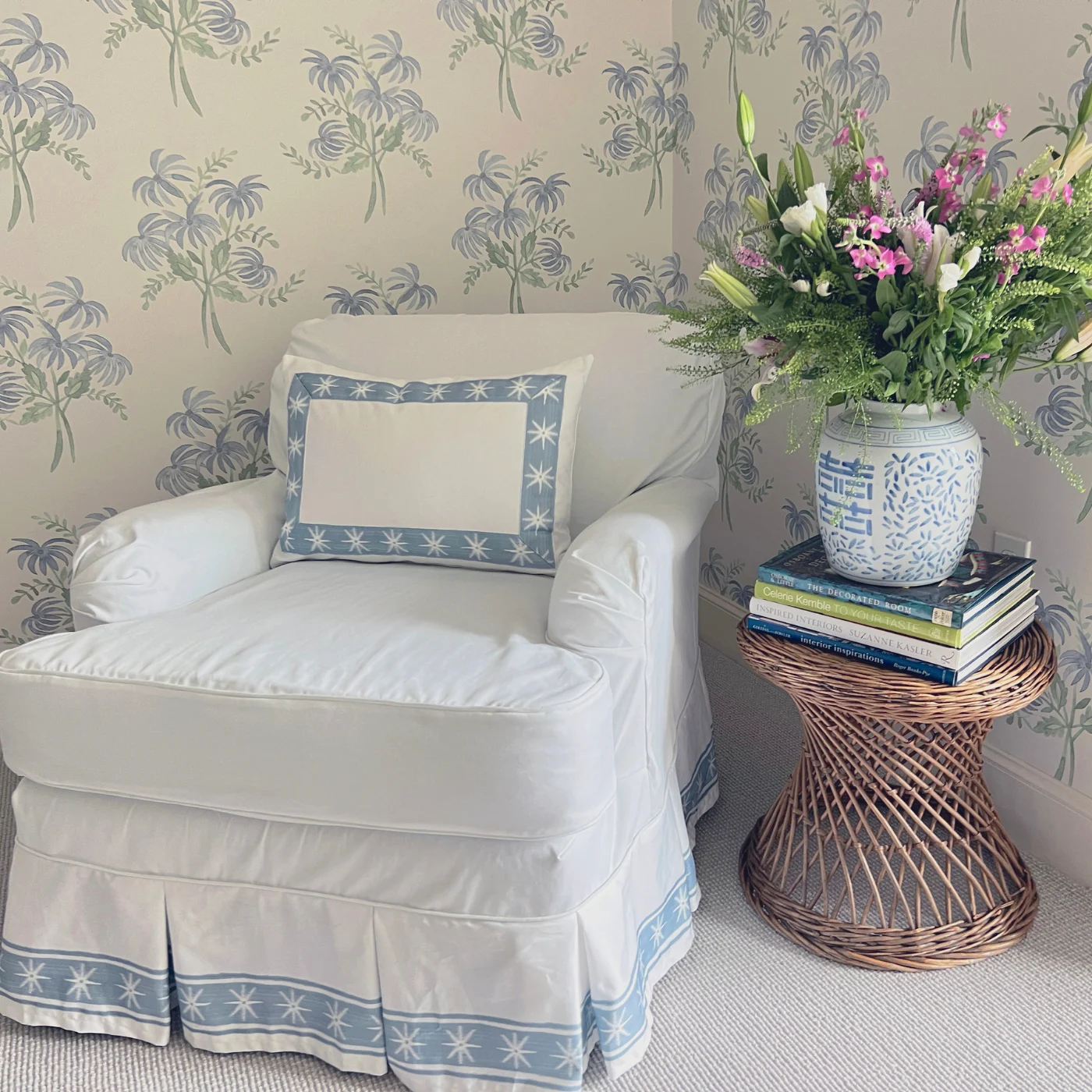A white pillow with blue fabric trim around the edges with white stars sits on a chair covered by a white slipcover with the same fabric trim at the bottom of the chair’s dust ruffles. The chairs sits in a living room corner in front of a wall covered in