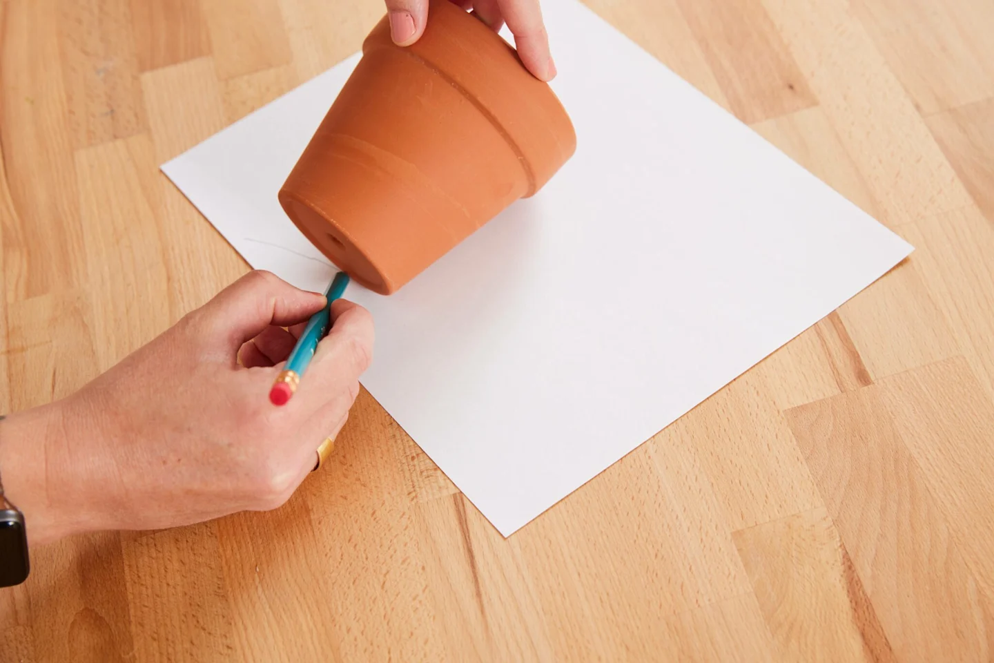 Alexa uses a pencil and a piece of paper to create a paper template for her fabric-covered flower pot. She is rolling the surface of the flower pot lengthwise across the paper. As she moves to the right, this creates an arc-like shape to be used as a temp
