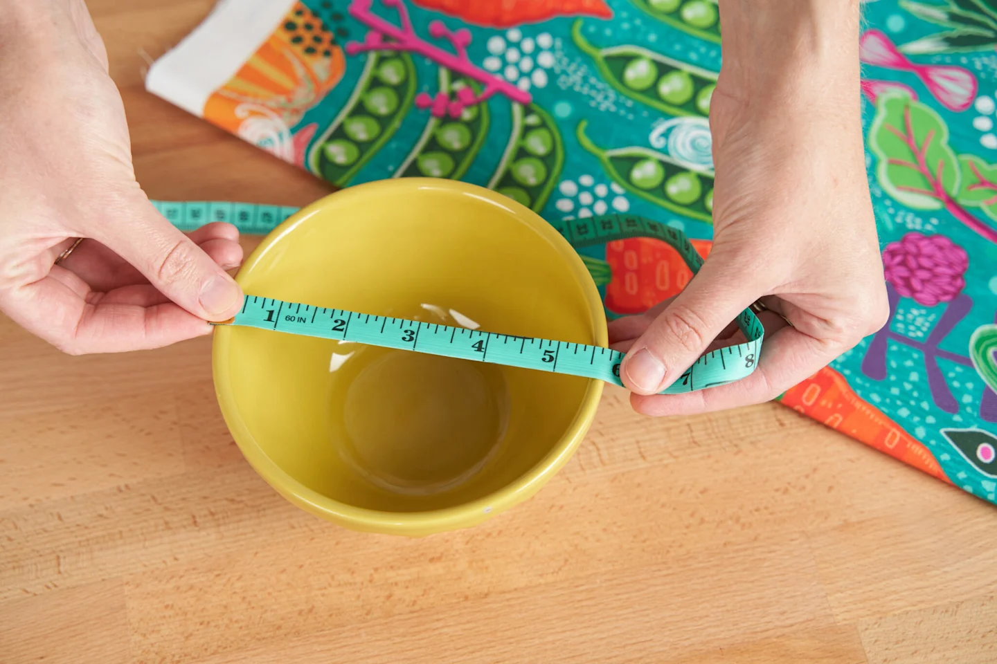 A photo of a person measuring the diameter of a yellow ceramic bowl that is sitting on a wooden table with a light green measuring tape. Only their hands are visible. A piece of a fabric featuring a design with tomatoes, cabbage and onions on a teal backg