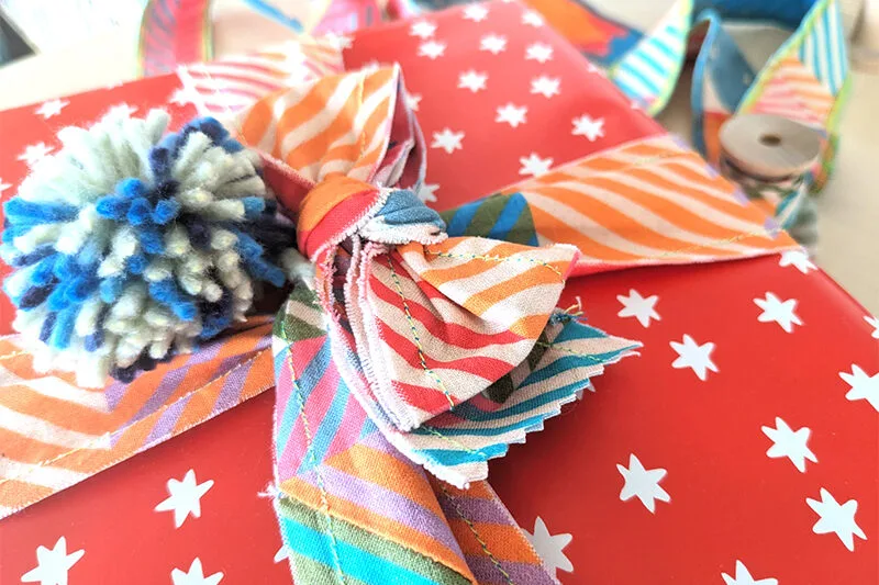 Close up of the top of a present wrapped in red wrapping paper with small white stars. Ribbon wrapped around the present has been made out of a 2”-wide fabric strips sewn together. The ribbon features a design with small squares of coordinating colors in 