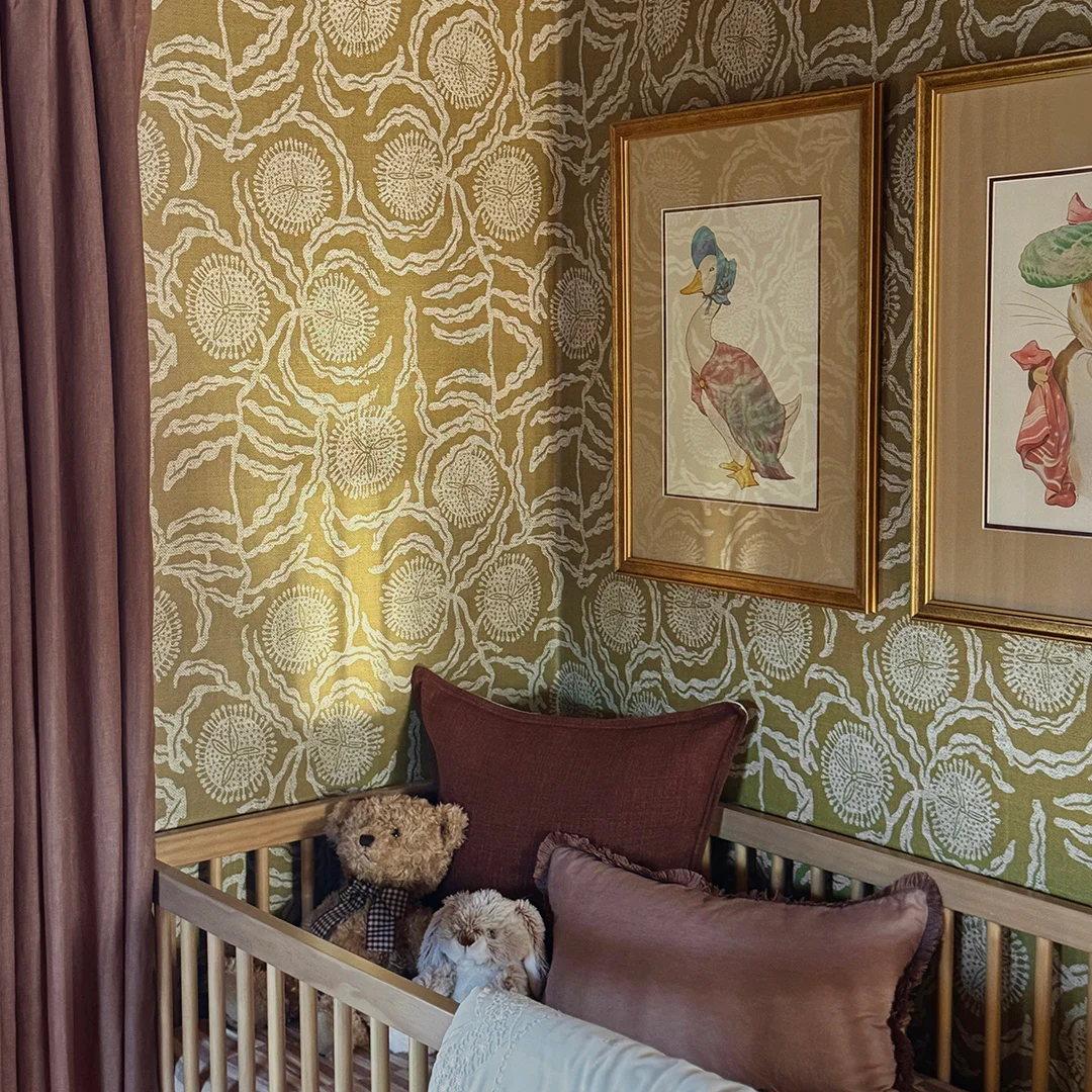 Nursery corner with olive green botanical wallpaper, mauve curtains, framed animal artwork, and a wooden crib styled with plush toys and pillows.