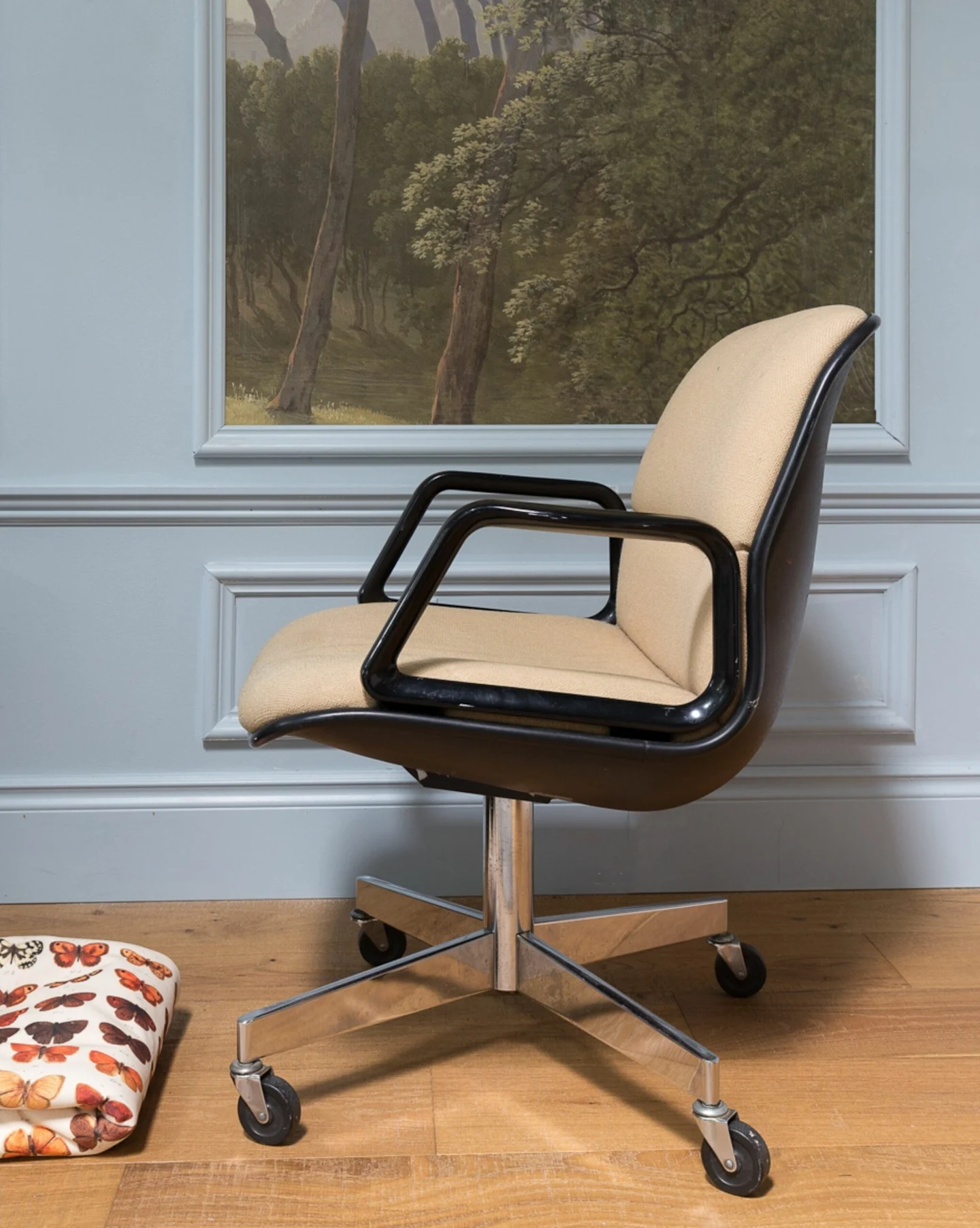 Image of a beige drop-seat chair in front of a light blue wall and on a light wooden floor, the photo is taken from the side.